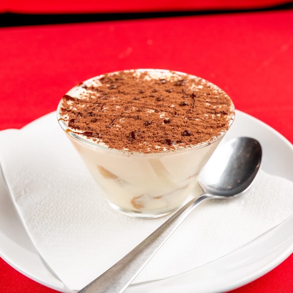 Tiramisu dessert in a glass, topped with cocoa, on a white plate with a spoon, on a red tablecloth.
