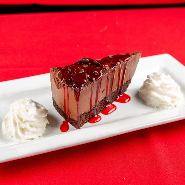 Decadent chocolate cheesecake slice topped with red berry sauce and two dollops of whipped cream.