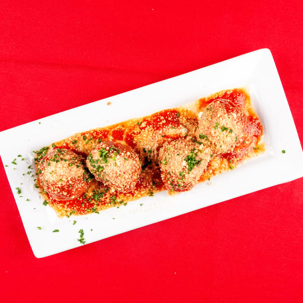 Five meatballs with red sauce, grated cheese, and parsley on a white plate over a red tablecloth.
