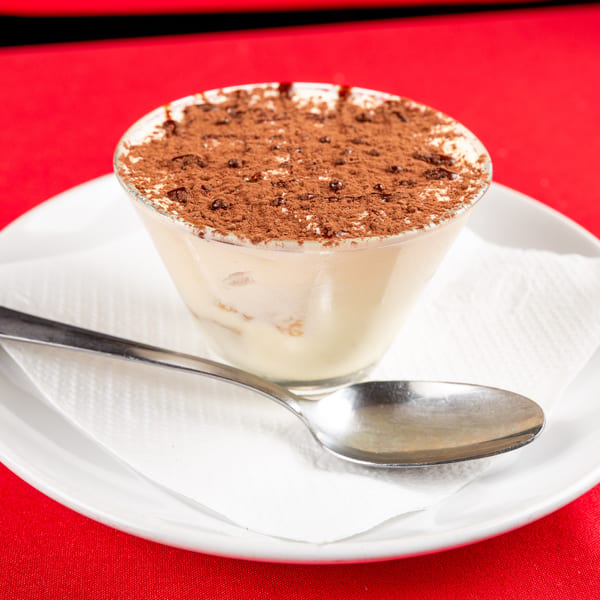 Tiramisu in a glass, topped with cocoa powder, on a white plate with a spoon and napkin.