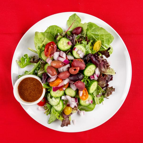 Mixed green salad with cucumbers, tomatoes, red onions, olives, and a side of dressing.