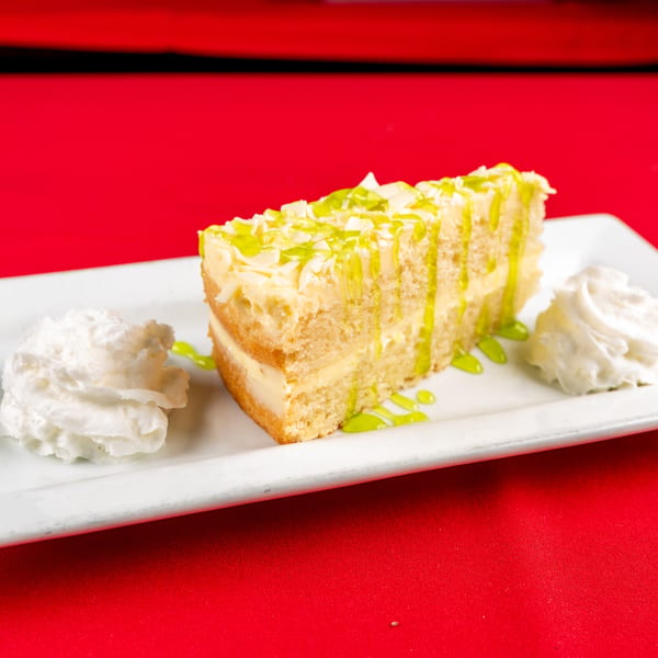 Slice of cake with green drizzle, white toppings, and two dollops of whipped cream on a white plate.