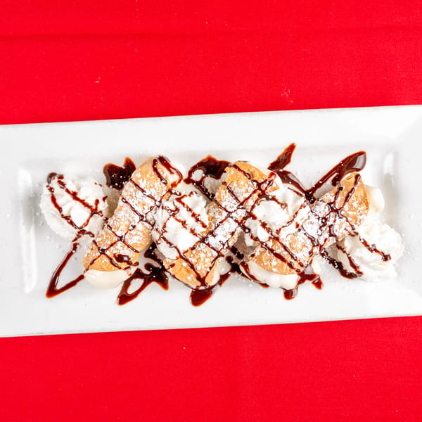 Three cannolis with whipped cream, chocolate drizzle, and powdered sugar on a white rectangular plate.