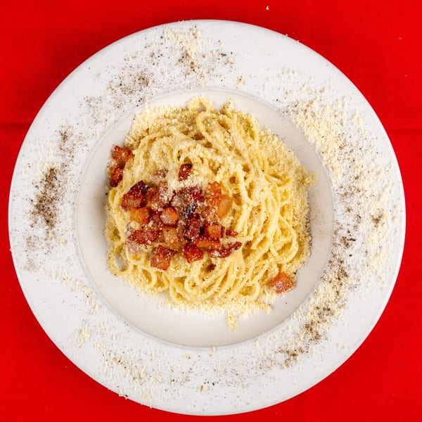 Plate of spaghetti carbonara with bacon and cheese, garnished with pepper, on a red background.