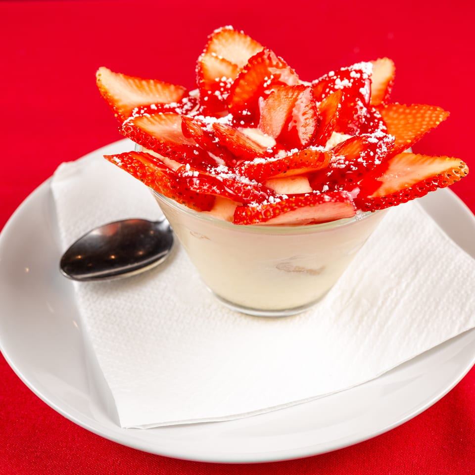 Creamy dessert in a glass, topped with sliced strawberries and powdered sugar, on a white plate.